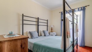 Una habitación con cama, cómoda de madera, mesita de noche, lámpara y ventana con cortinas.