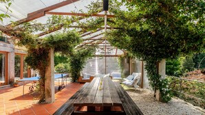 Un patio cubierto con una mesa y sillas de madera, rodeado de vegetación y una pérgola.