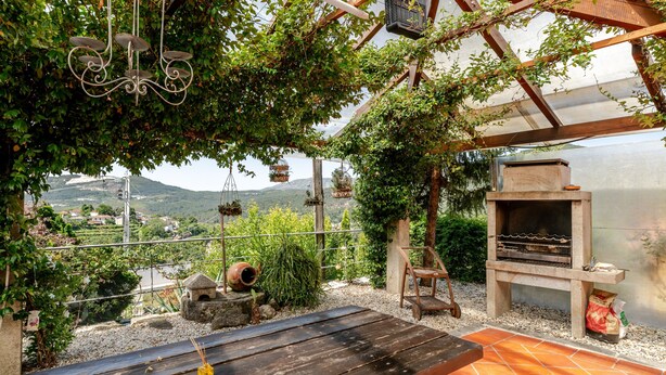A patio with a wooden table, hanging baskets, a grill, and a view of a mountainous landscape.