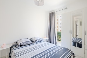 A bedroom with a bed, a bedside table, a mirror, and a window with curtains.