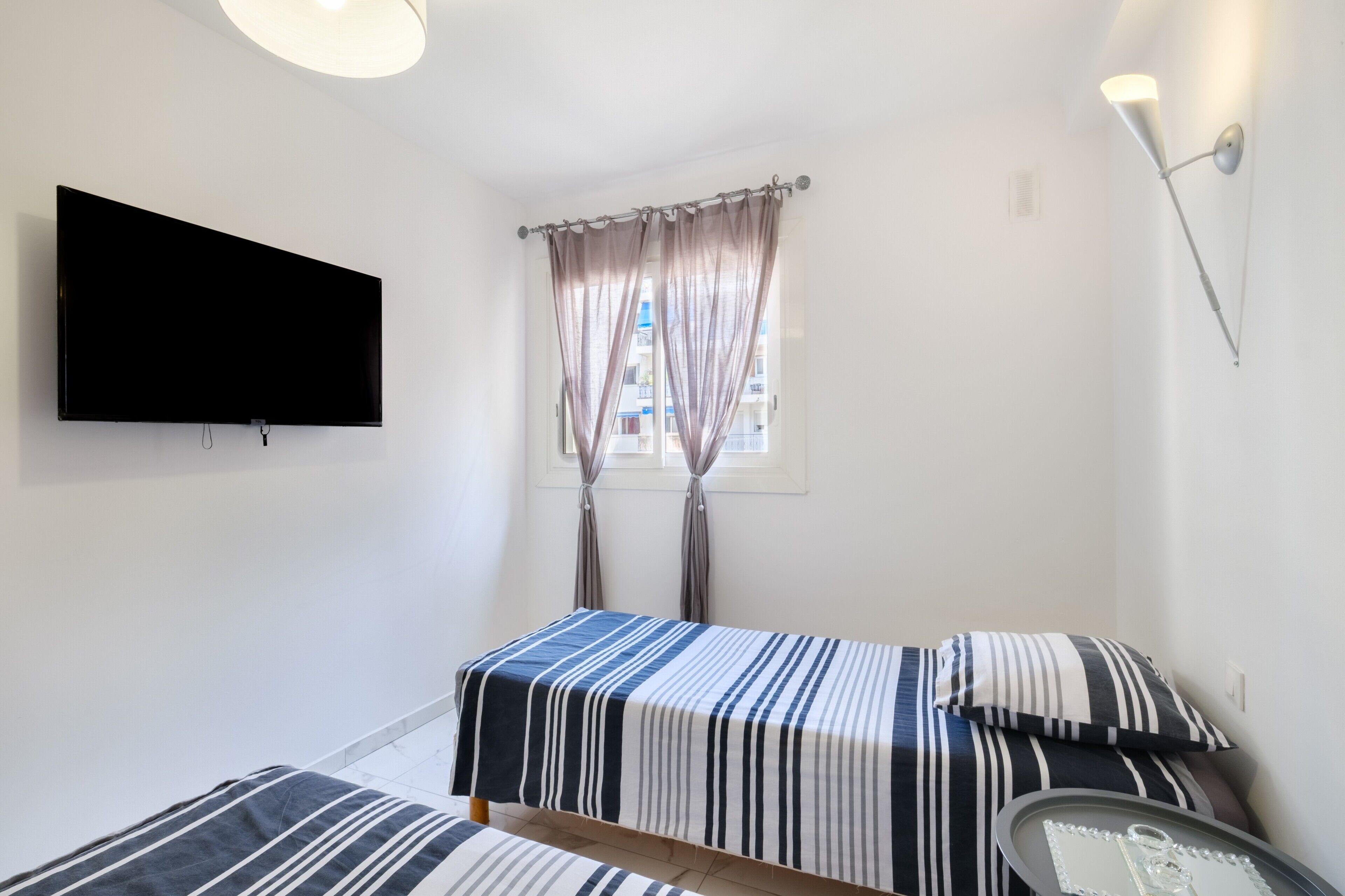 Una habitació amb televisió de pantalla plana penjada a la paret, dos llits amb roba de llit a ratlles, una finestra amb cortines i una làmpada penjada a la paret.
