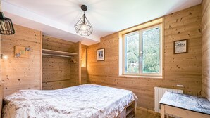 Ein Zimmer mit Holzwandverkleidung, ausgestattet mit einem Bett, einem Tisch, einer Hängeleuchte und einem Fenster mit Blick auf Grünfläche.