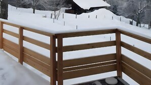 Eine Holzterrasse mit verschneiter Landschaft, darunter Bäume und ein Haus im Hintergrund.