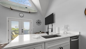 A modern kitchen with a large island, a mounted surfboard, a clock, and a TV on the wall. - Clean & Updated Just Steps from the Beach. Dogs Welcome. Grill & Beach Gear (Surf City)