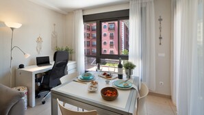 Een moderne hotelkamer met een eettafel gedekt voor een maaltijd, een laptop op een bureau en uitzicht op een rood gebouw door het raam.