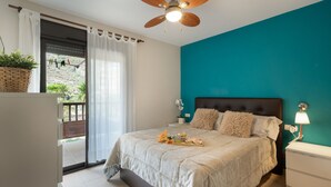 Een slaapkamer met een bed, een nachtkastje, een lamp, een plafondventilator en uitzicht op een balkon met planten en een drooglijn.