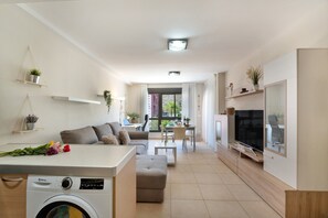 A compact living space with a washing machine, a sofa, a dining area, and a TV.