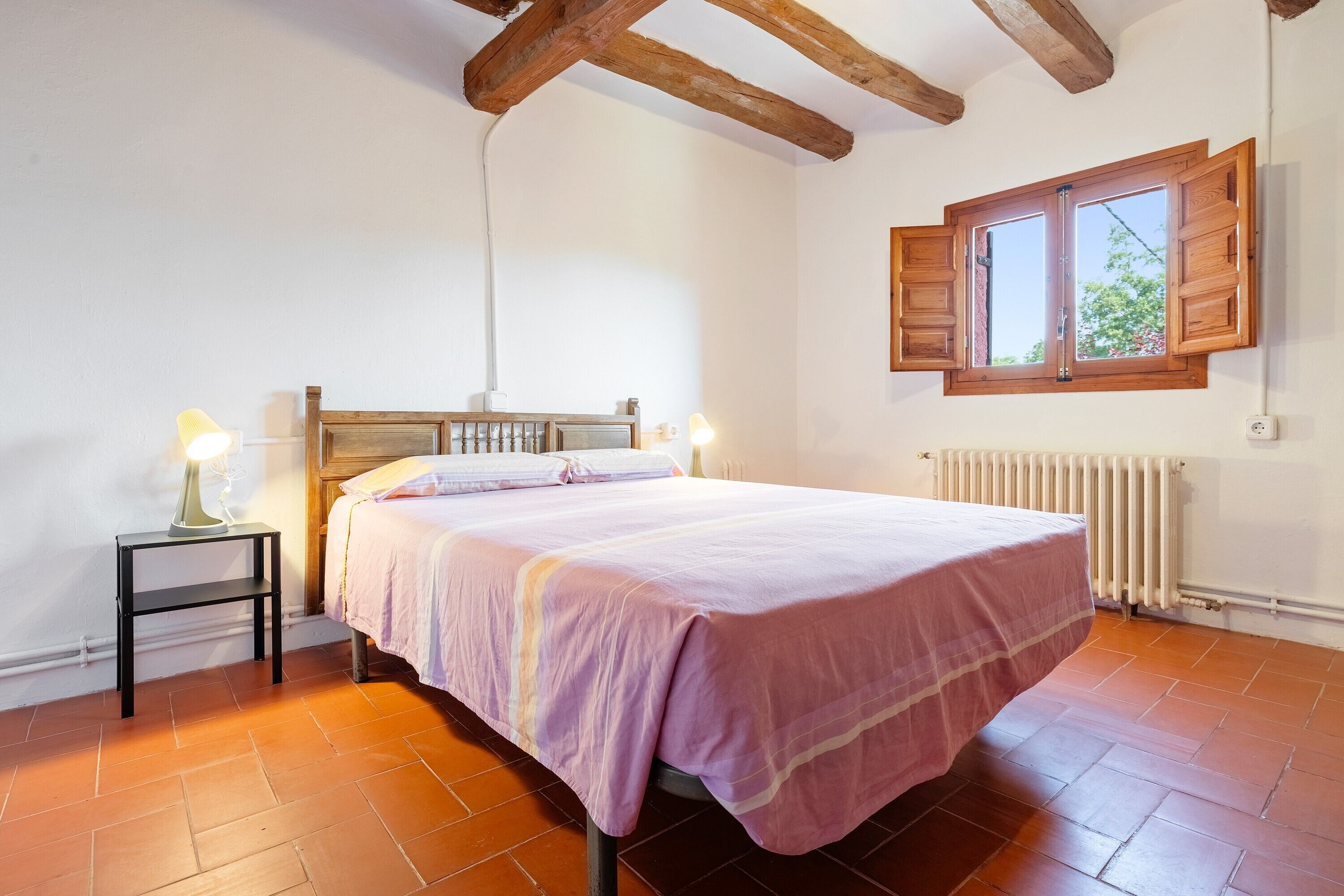 Un dormitorio con cama, mesita de noche, ventana con contraventanas y radiador.