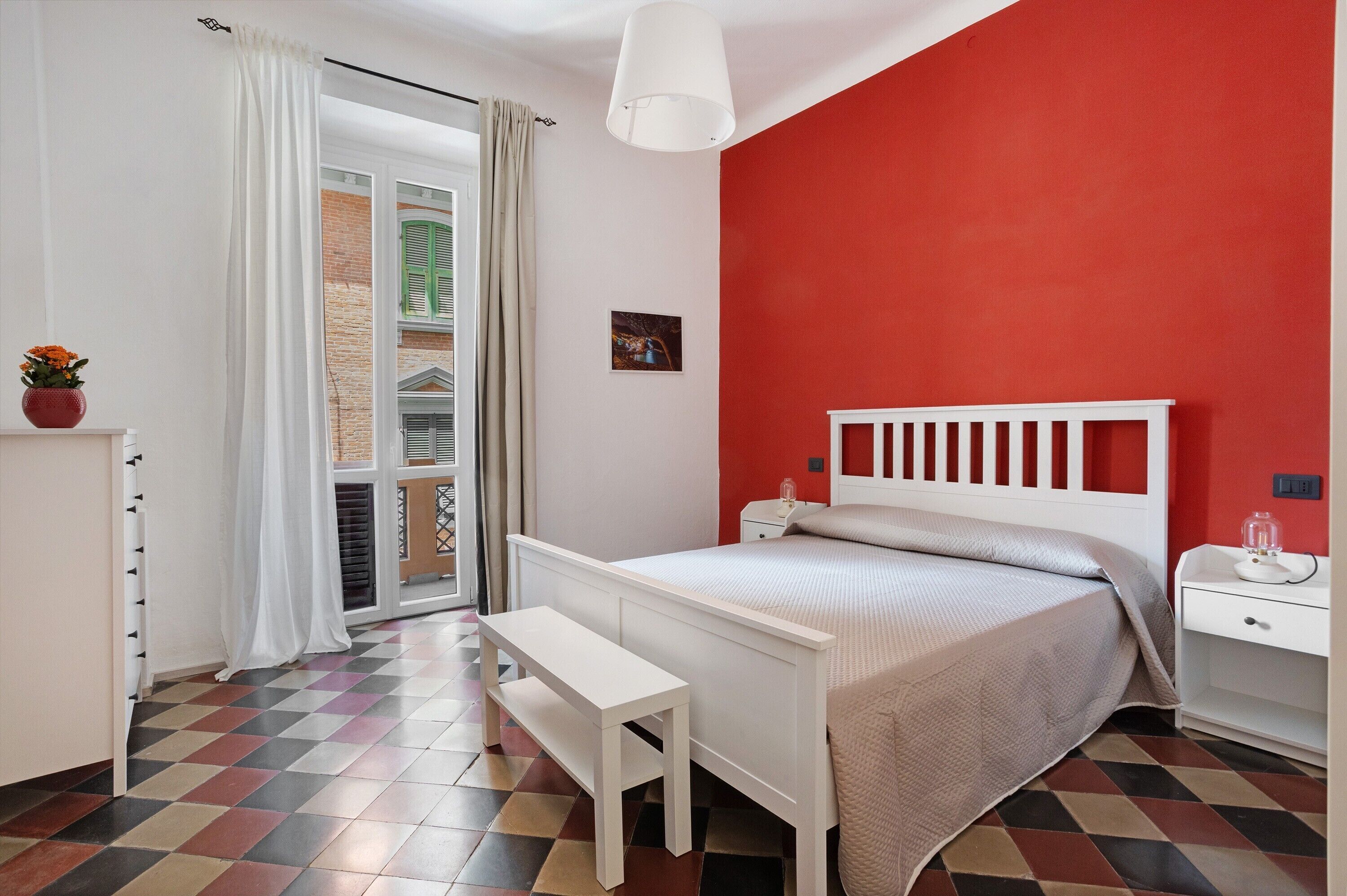 Ein Schlafzimmer mit kariertem Boden, einem weißen Bett, einem Nachttisch, einer Bank und einer roten Wand.
