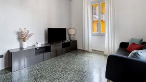 A modern living room with a flat-screen TV, a console table, a vase with dried flowers, a sofa with cushions, and a window with curtains. - La Casa Di Licia - Apartment in La Spezia (La Spezia)