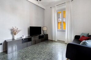 A modern living room with a flat-screen TV, a console table, a vase with dried flowers, a sofa with cushions, and a window with curtains.