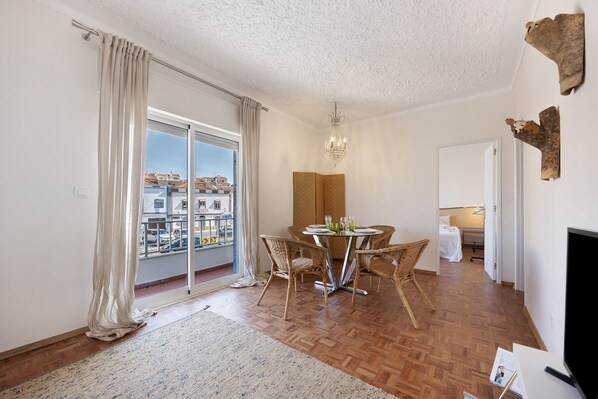 A room with a dining table, chairs, a chandelier, a sliding glass door, and a bed in the background.