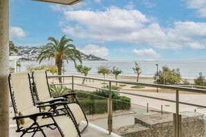 A balcony with two lounge chairs, a view of the sea, and a distant cityscape.