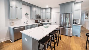 A modern kitchen with grey cabinets, a stainless steel refrigerator, a microwave, and a large island with bar stools. - Swans Rest Townhouse, Close to Beach & Restaurants (Cape Charles)