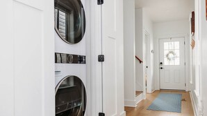 A laundry room with two large front-loading washing machines, a white door, a wooden staircase, and a blue patterned rug. - Swans Rest Townhouse, Close to Beach & Restaurants (Cape Charles)