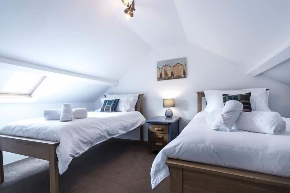 Quarto de hotel com duas camas, uma mesa pequena, uma mesinha de cabeceira, uma luminária e um quadro na parede.