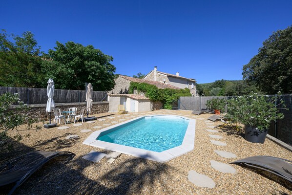 A swimming pool with a stone border, surrounded by pebbles, with chairs and umbrellas.