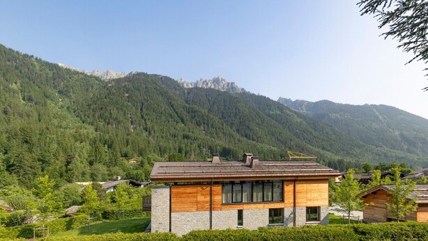 Una casa moderna de dos pisos con fachada de piedra y niveles superiores de madera, rodeada de abundante vegetación y montañas.