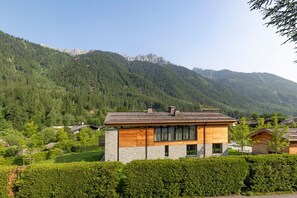 Une maison moderne à deux étages, dotée d’une façade en pierre et d’étages supérieurs en bois, entourée d’une végétation luxuriante et de montagnes.