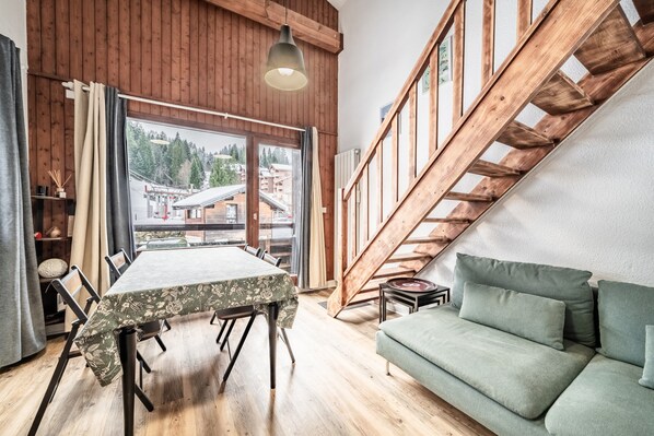 Une pièce comprenant une table à manger, un canapé, un escalier en bois et une grande fenêtre donnant sur des immeubles et des arbres.