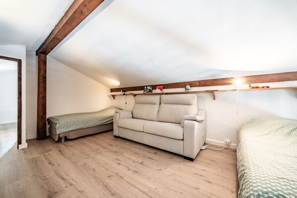 A room with a wooden ceiling, a beige sofa, a small bed, and a shelf with books and a lamp.