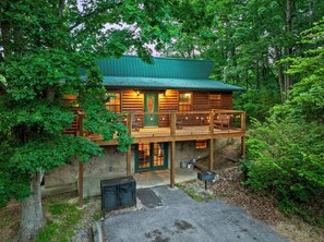 Une cabane en rondins, avec des fondations en pierre, une terrasse en bois et un toit végétalisé, entourée d’une forêt dense.