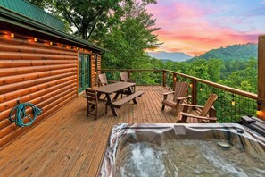 Une terrasse en bois avec un jacuzzi, une table de pique-nique et des chaises, offrant une vue sur un paysage boisé.