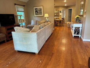 A living room with a sofa, a TV, a dining area with chairs and a table, and a hallway leading to another room. - Walk to beach in this charming home with ac and wifi (Riverhead)