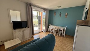 A living room with a blue sofa, a TV on a white cabinet, a dining table with chairs, and a kitchen area with a fridge. - 226 - Comfortable apartment in a residence with swimming pool in Verdon sur Mer (LE VERDON SUR MER)