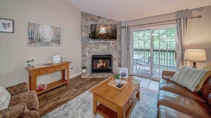 A cozy living room with a stone fireplace, a wooden coffee table, a leather sofa, and a large window with a view of greenery. - LOCATION!!  Only 1 mile to downtown Helen. 2 Bedroom, 2 Bath. Gated Community. (Helen)