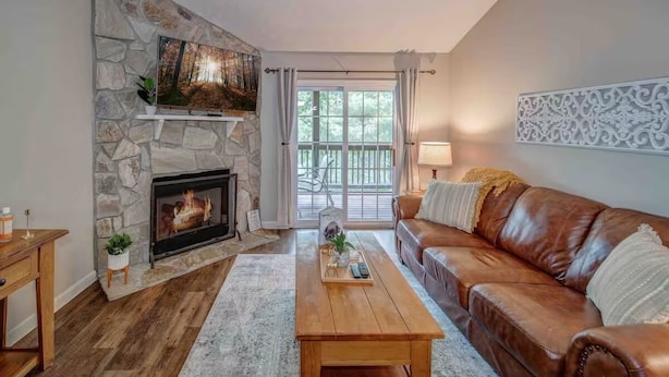 A living room with a stone fireplace, a leather sofa, a wooden coffee table, and a large window with curtains. - LOCATION!!  Only 1 mile to downtown Helen. 2 Bedroom, 2 Bath. Gated Community. (Helen)