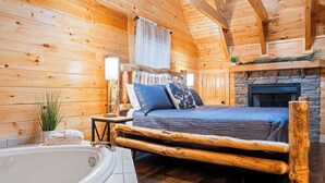 A log cabin bedroom with a large bed, a stone fireplace, and a bathtub.