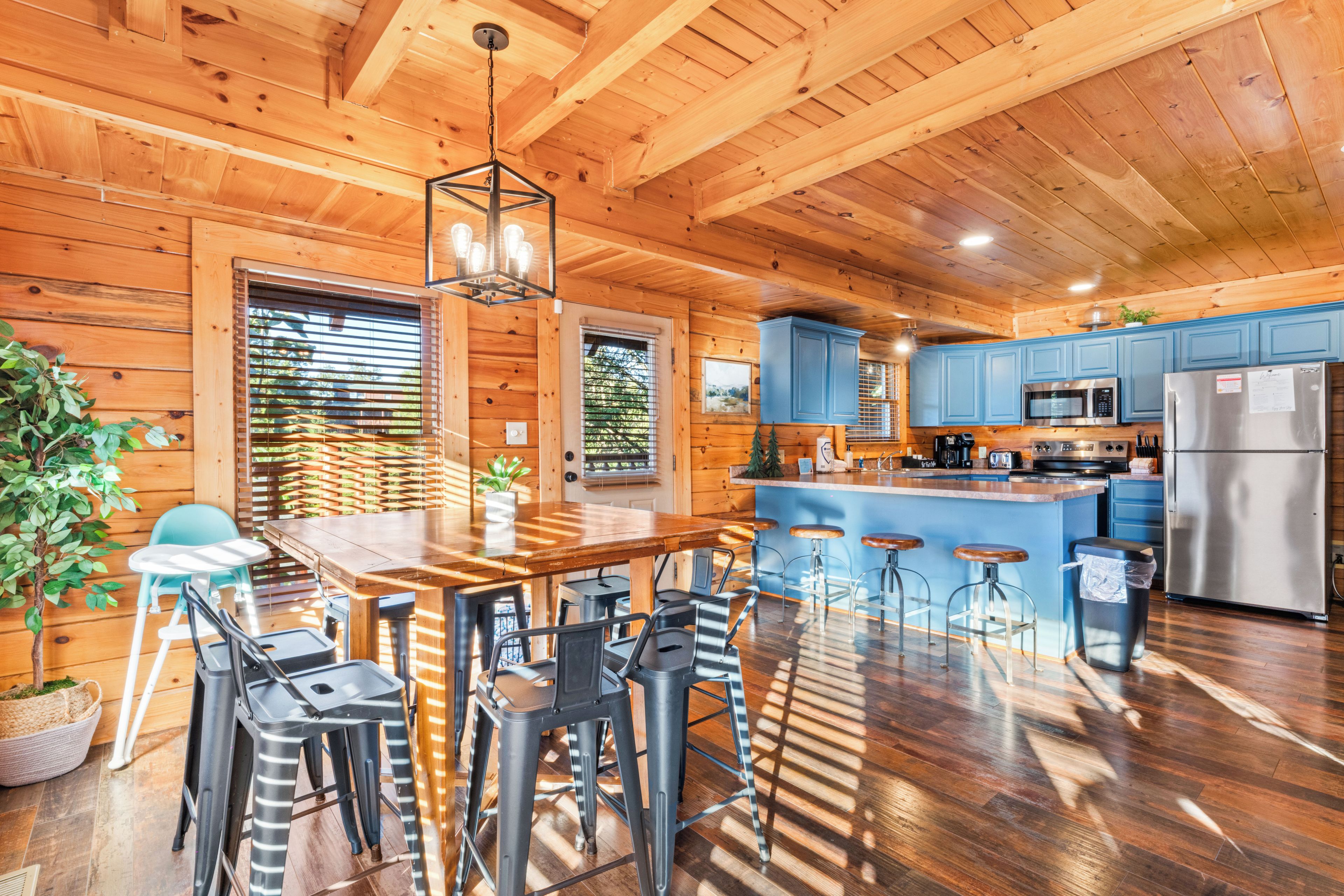 A wooden cabin interior with a central dining area, bar stools, a kitchen with blue cabinets, and a stainless steel refrigerator.
