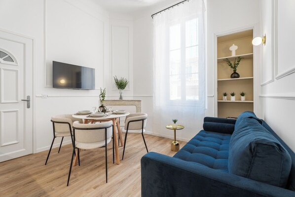 Ein modernes Wohnzimmer mit einem blauen Sofa, einem Esstisch mit Stühlen, einem an der Wand montierten Fernseher und einem Fenster mit durchsichtigen Vorhängen.