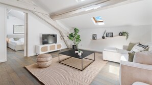 A modern loft bedroom with a sloped ceiling, skylights, a large bed, a TV, and a cozy seating area with a coffee table and plants.