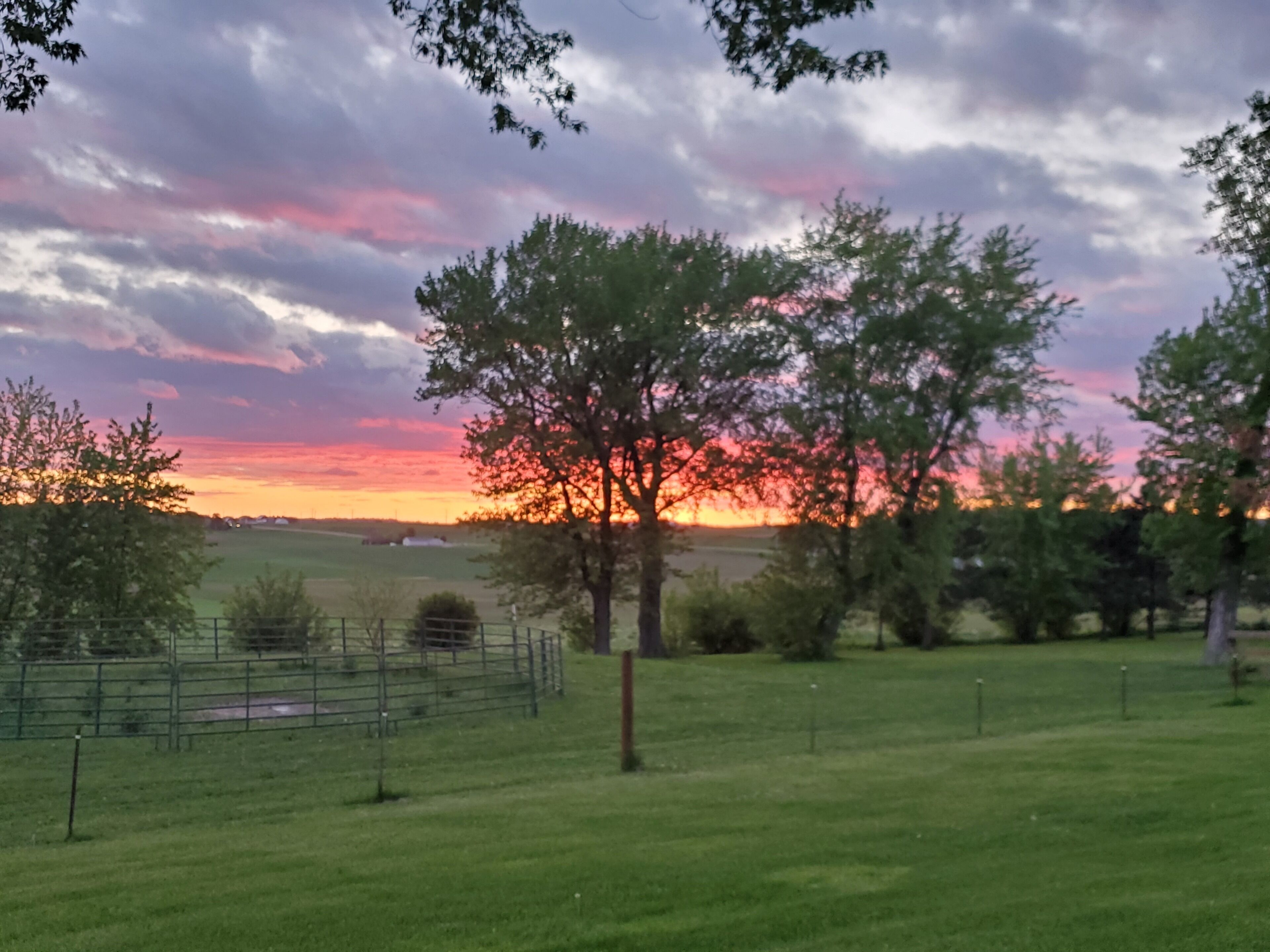 Ländliche Landschaft mit Sonnenuntergang, offenen Feldern und einem eingezäunten Bereich.