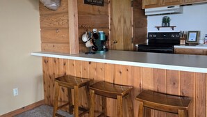Une cuisine avec des lambris en bois, une tête de cerf accrochée au mur, un bar avec des tabourets et une pancarte sur laquelle on peut lire : « LA MAISON, C’EST L’ENDROIT OÙ VOUS ACCROCHEZ VOS TROPHÉES DE CHASSE. »