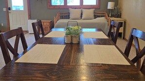 Un coin repas avec une table et des chaises en bois, une fenêtre donnant sur les arbres, un lit avec un oreiller à carreaux et une lampe sur une table de chevet.