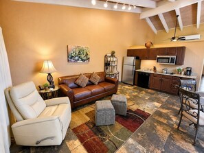 Een woonkamer met een bank, fauteuil en voetenbankje, een keuken met apparatuur, en een plafondventilator.