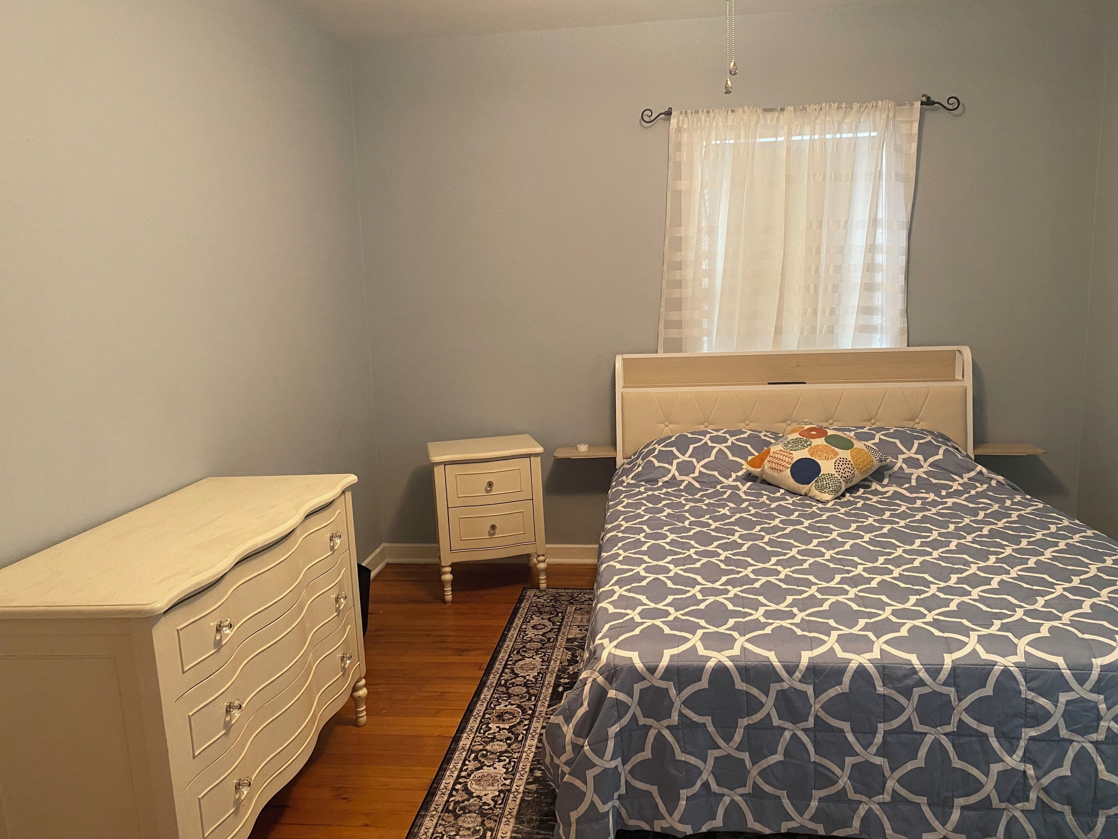 Een slaapkamer met een bed, nachtkastje, dressoir en een raam met gordijnen.