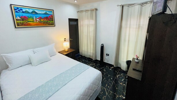 A hotel room with a bed, a painting, a desk with a lamp, a TV, and curtains. - hostal bode de lago rooms  #3 (Panajachel)