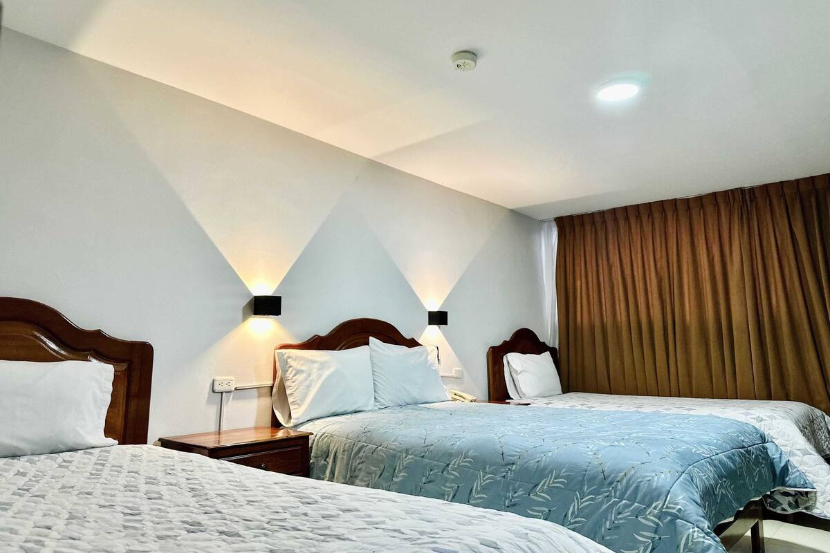 A hotel room with two beds, a wooden headboard, a nightstand with a lamp, and a window with curtains.