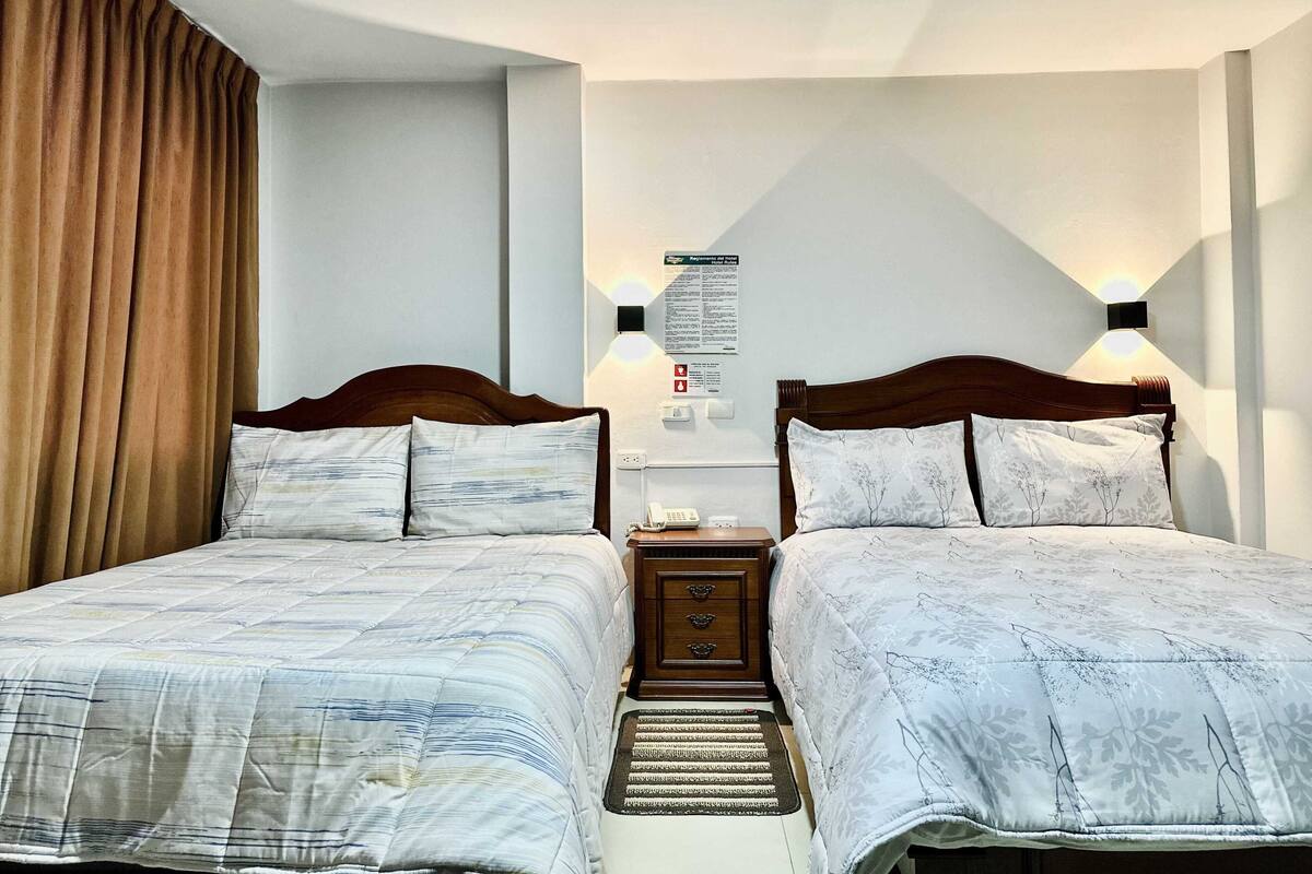 A double room with two beds, a nightstand, a wall-mounted light, and a bulletin board with information.