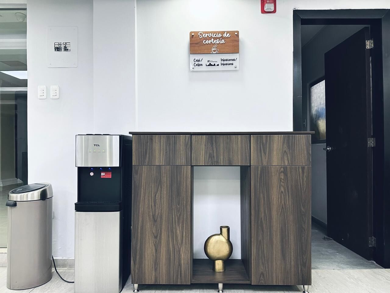 Een modern receptiebalie met een koffieapparaat, een prullenbak en een houten kastje met een vaas.