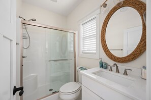 Une salle de bain moderne dotée d’une douche à parois de verre, de toilettes blanches, d’un lavabo blanc avec deux robinets et d’un grand miroir avec un cadre tressé.