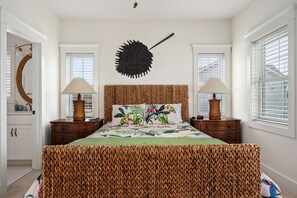 Une chambre à coucher avec une tête de lit en osier, deux tables de chevet avec des lampes, une couvre-lit à motifs floraux et un grand élément décoratif mural.