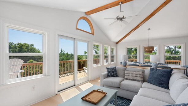 A spacious living room with a large sofa, a coffee table, and a ceiling fan. There are multiple windows providing a view of the outdoor deck.