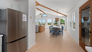 A modern kitchen with a stainless steel refrigerator, wooden beams, a dining area with blue chairs, and large windows offering views of the outdoors.