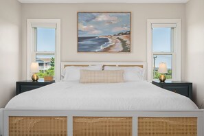 Quarto com uma cama grande, duas mesas de cabeceira com luminárias, um quadro de uma praia e janelas com vista para o verde.