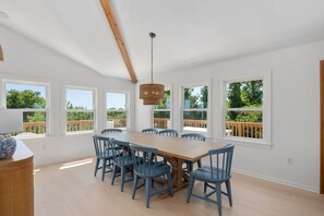 Ein geräumiger Essbereich mit einem großen Holztisch und blauen Stühlen, großen Fenstern mit Blick auf Grünflächen sowie einer hängenden Korbschwinge als Lampenleuchte.